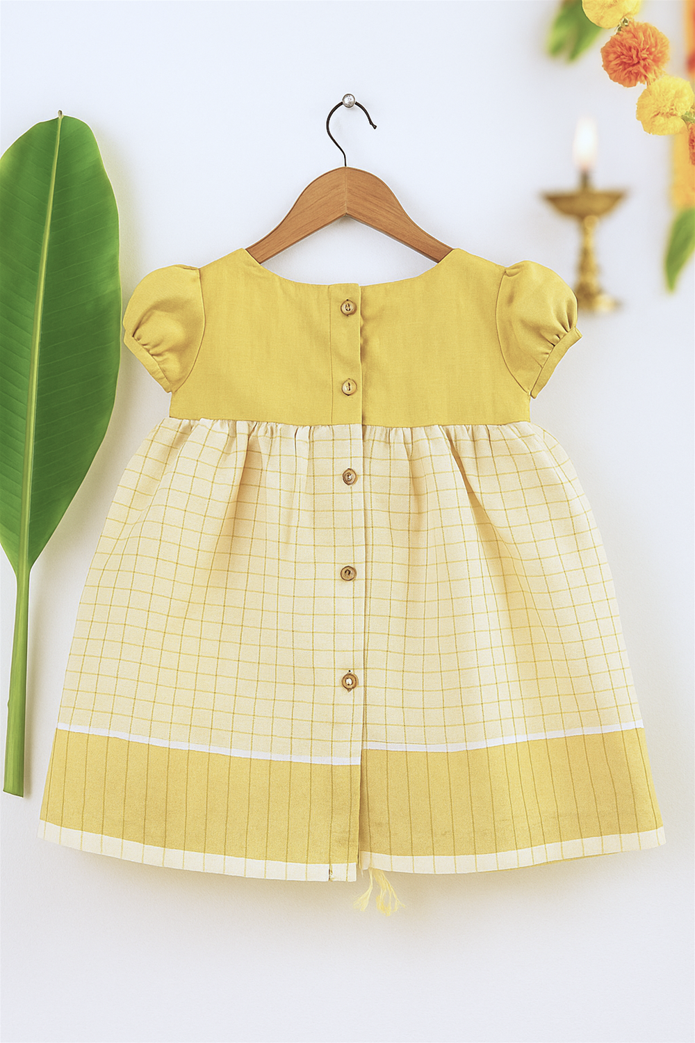 A yellow and cream checkered baby dress with button detailing on the back is displayed on a wooden hanger. The backdrop includes a banana leaf and festive elements, enhancing its traditional charm.
