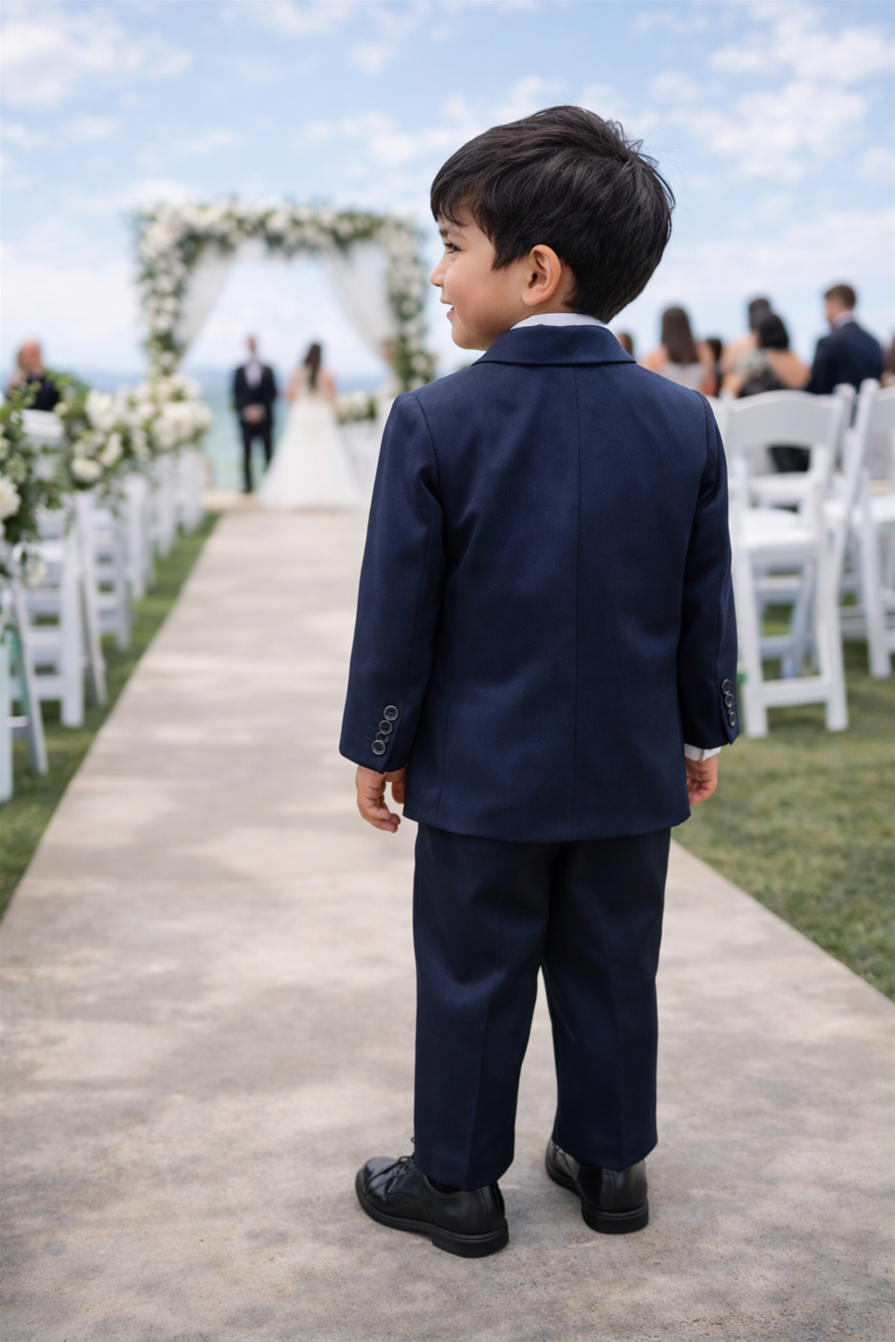 Toddler boy wearing formal suit walking outdoors on grass, seen from behind at a garden wedding ceremony setup.