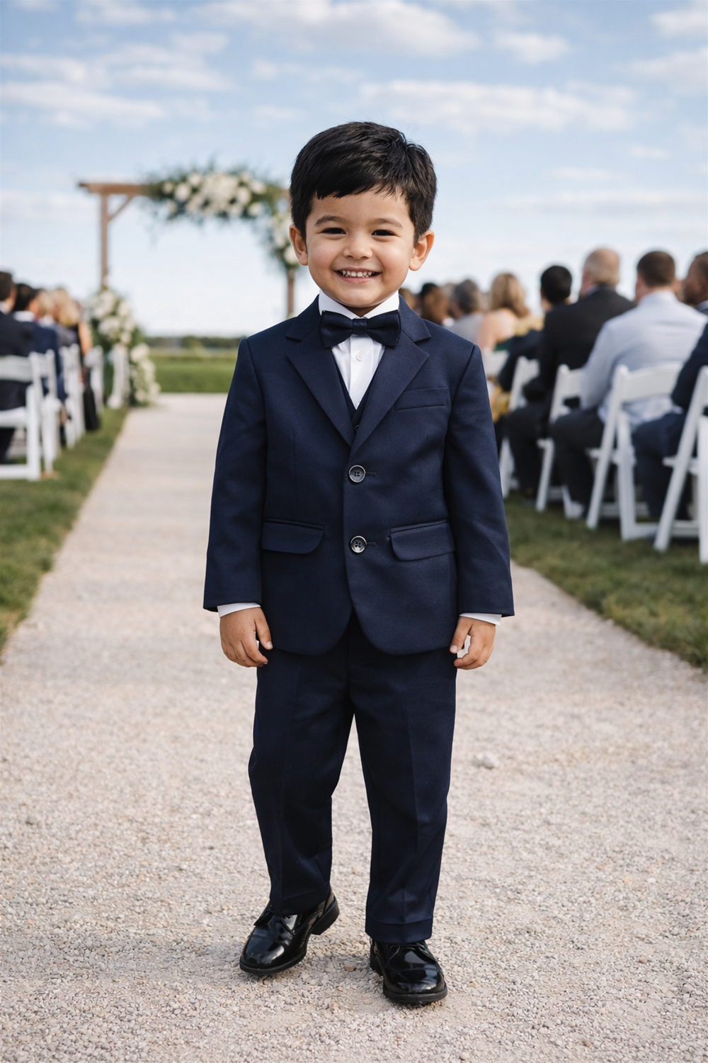 Smiling toddler boy in suit and bow tie standing outdoors at wedding aisle with floral arches and guests.