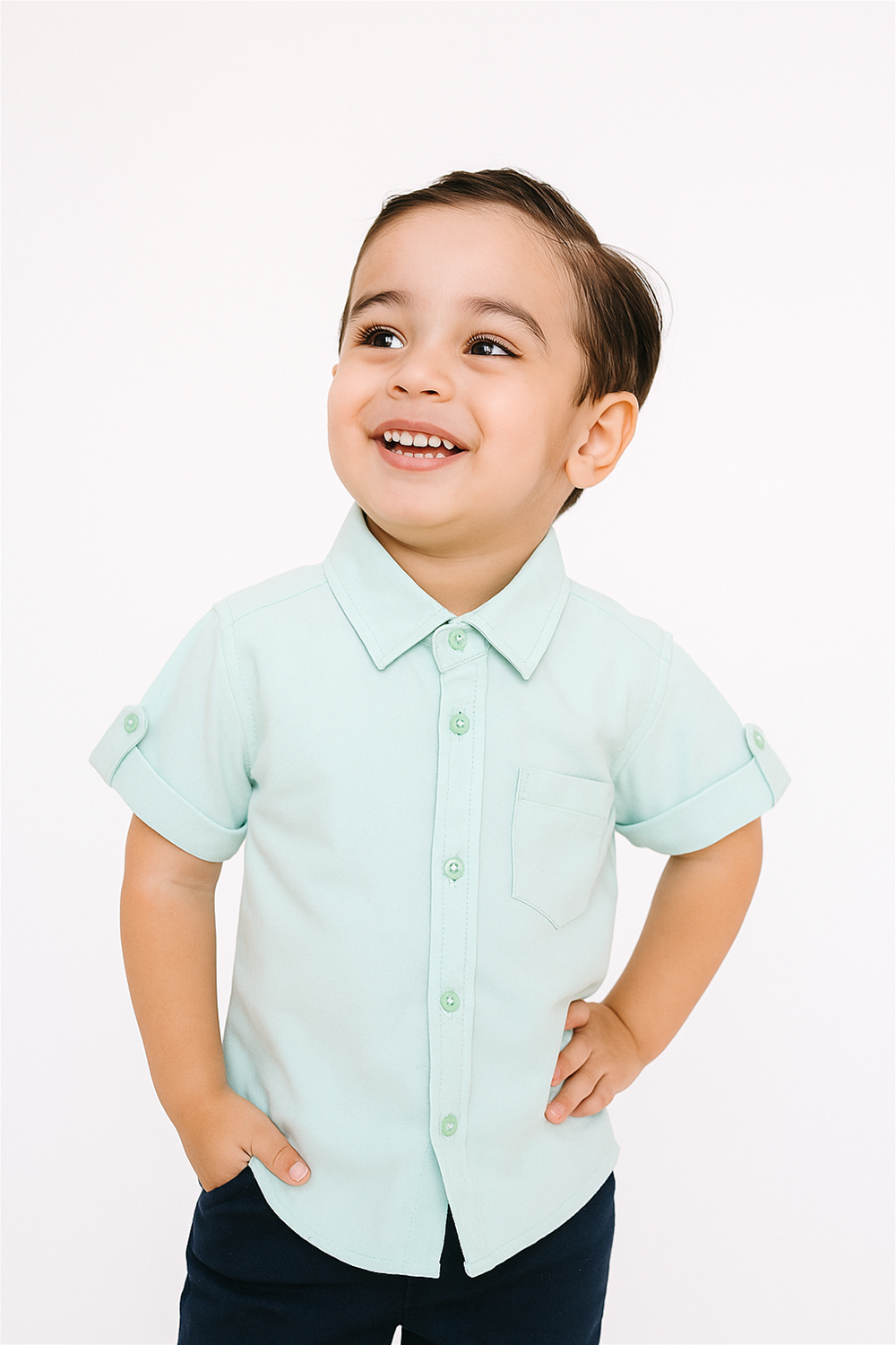 Front view of a cheerful boy wearing a mint green half sleeves Chinese collar shirt with turned-up cuffs and a chest pocket. The shirt is paired with dark trousers and photographed against a plain white background.