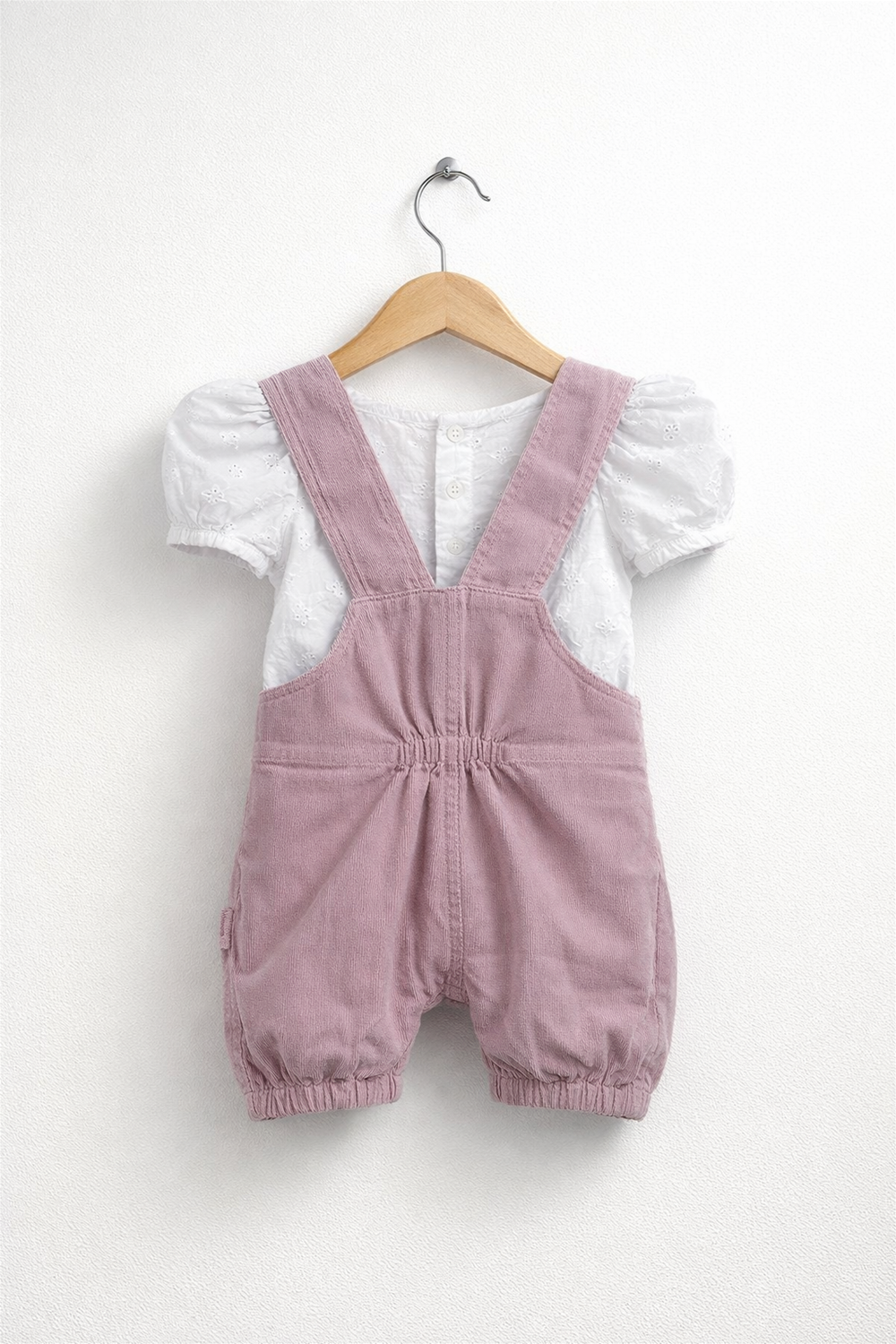 Onion Pink baby dungaree with ruffled straps paired with white embroidered top, neatly displayed on a wooden hanger against wall.
