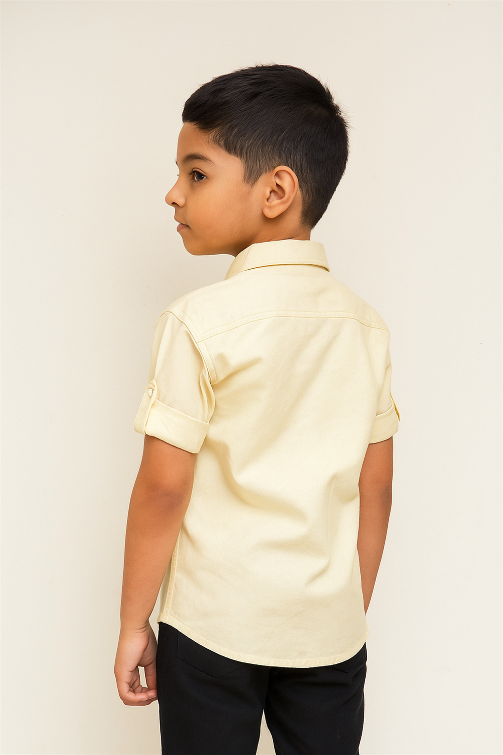 A young child stands confidently in a light yellow button-up shirt with rolled sleeves, paired with black pants against a white background.