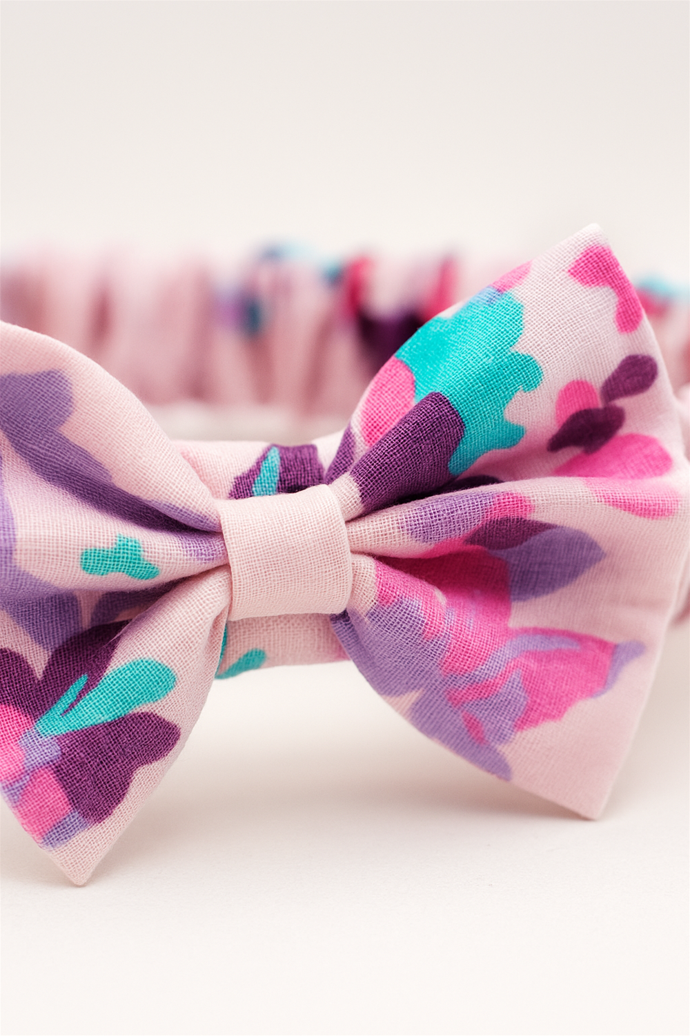 A pink elastic baby headband with a gathered fabric band, featuring a decorative bow in a pastel floral pattern with shades of purple, turquoise, and pink. The headband is laid flat against a white background, showcasing its design in full detail.