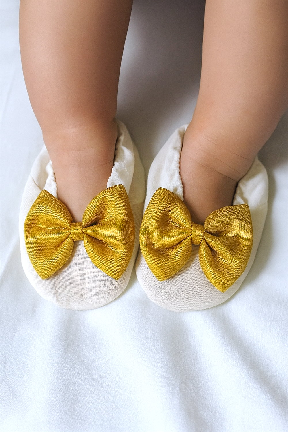Close-up of a baby’s feet in cream booties decorated with shiny golden bows. The shoes fit snugly and enhance the adorable appearance of the little one.