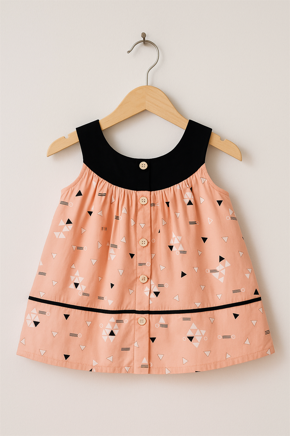 Peach sleeveless frock with black neckline and geometric print, displayed on a wooden hanger, buttons aligned along the center.