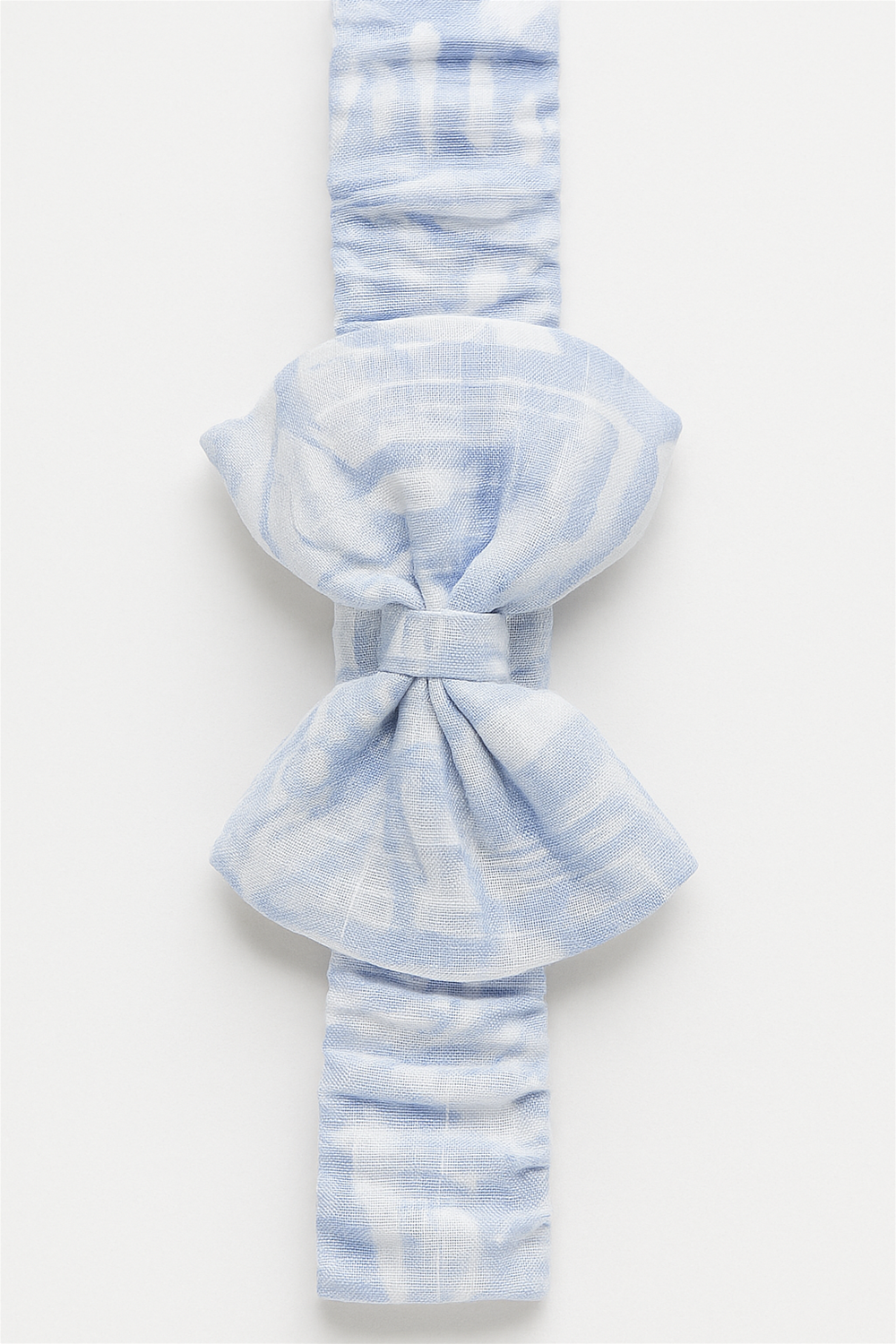 A soft, stretchy baby headband featuring a large bow in a light blue tie-dye pattern. The fabric is textured and ruched for comfort, designed to fit snugly while adding a charming touch to any baby outfit.