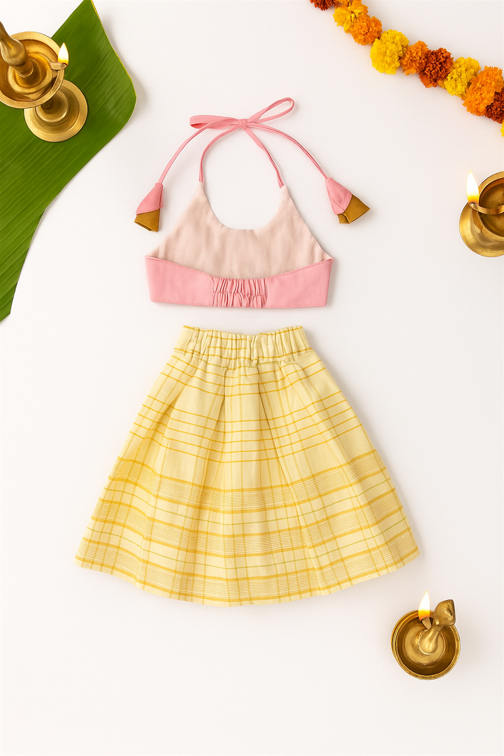 Flat-lay of a baby girl’s festive outfit with pink halter top, yellow checked skirt, decorative tassels, surrounded by lamps.