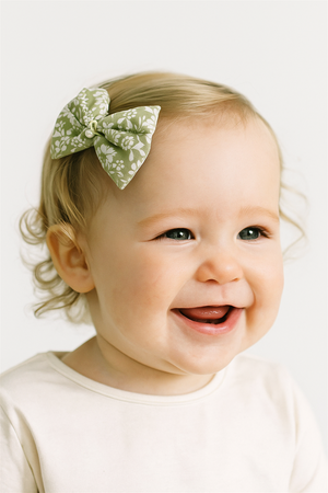 Smiling baby girl wearing green floral bow hair clip with pearl center, styled neatly on hair, captured in bright light.