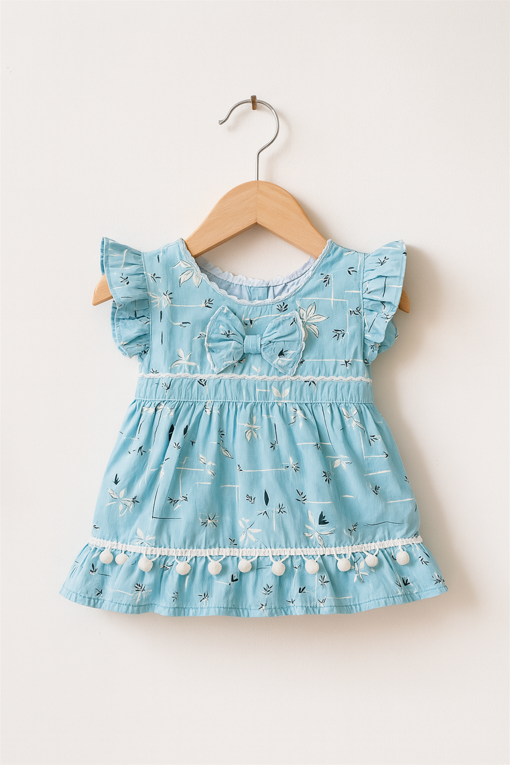 A charming sky blue baby dress with delicate floral prints hangs on a wooden hanger against a white wall. The dress features flutter sleeves, a center bow, and white pom-pom trim along the hem.