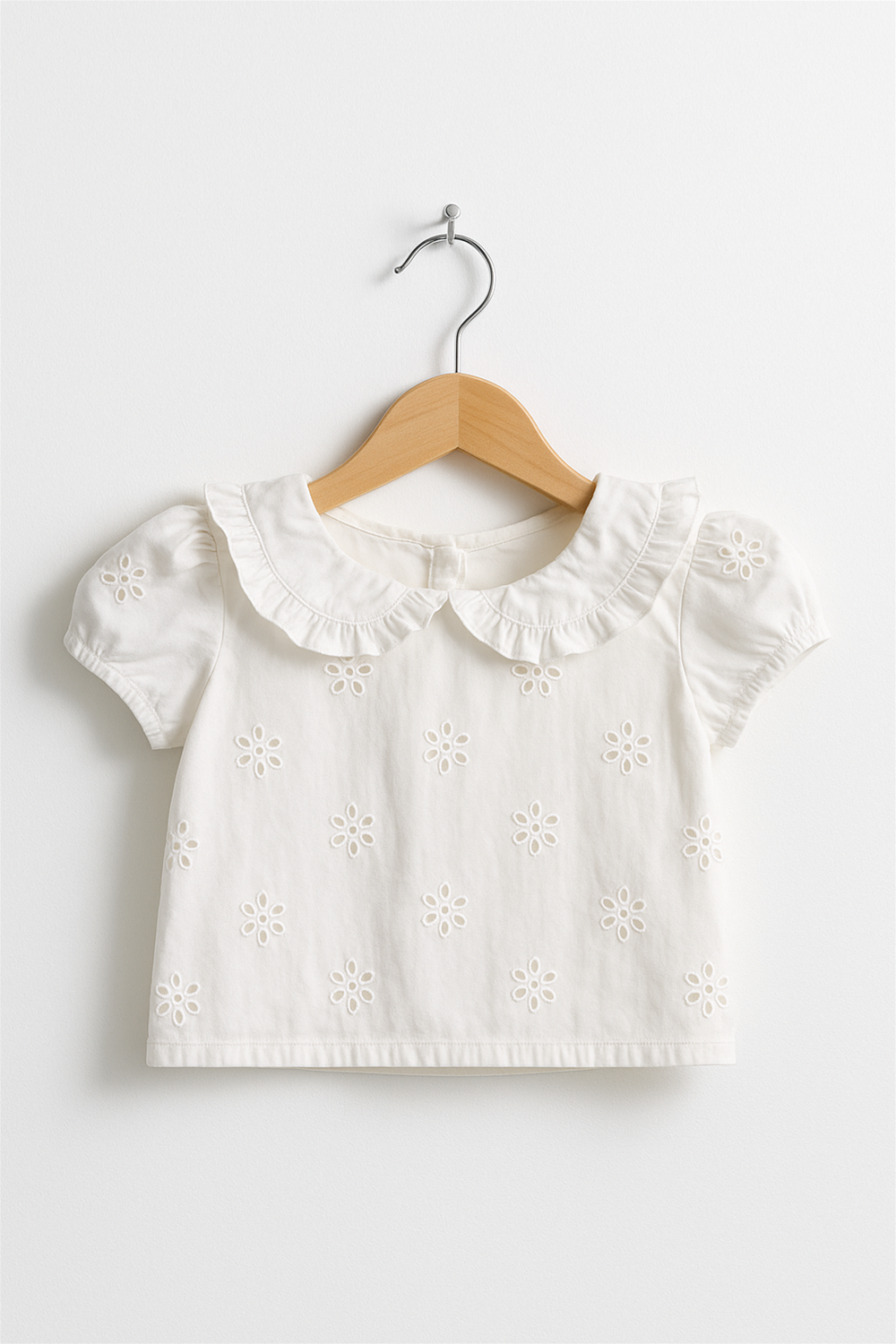 White toddler blouse with a ruffled Peter Pan collar, short puff sleeves, and all-over daisy eyelet embroidery. Displayed on a wooden hanger against a plain white wall.