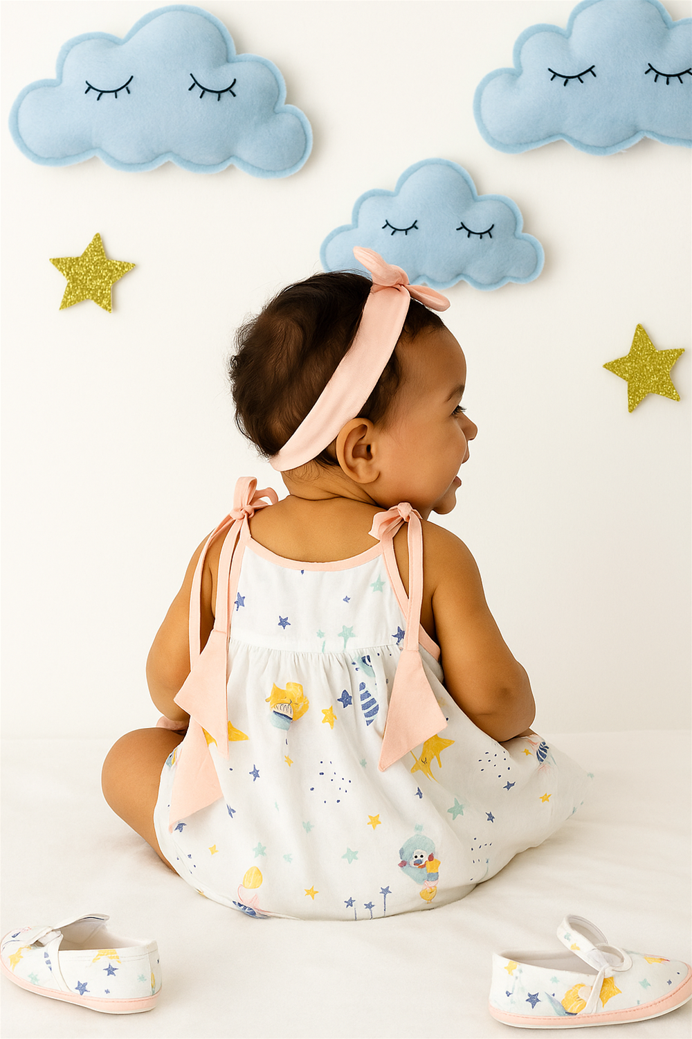 Adorable baby girl seated with her back facing the camera, wearing a white dress with colorful space-themed prints and peach bow-tie straps. The background features plush blue clouds and golden stars, creating a dreamy, celestial nursery vibe.