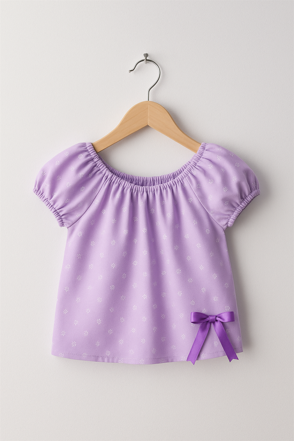 Lavender embroidered girls’ top with puff sleeves and gathered neckline, enhanced with a decorative satin bow detail near the hemline.