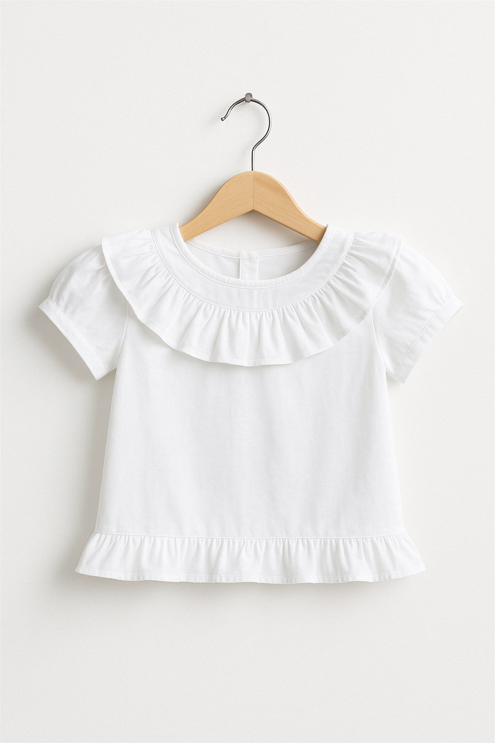 White cotton top with round neck, short sleeves, front ruffle design, and hem frill, displayed on a wooden hanger.