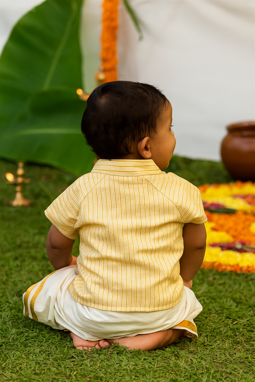 Baby boy sitting with back view, wearing yellow striped kurta and white dhoti, watching floral rangoli with festive decor behind.