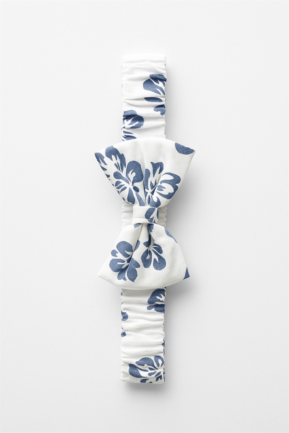 A white elastic headband featuring a navy-blue floral print, designed with a central bow for a stylish and charming look. Perfect as an accessory for babies and toddlers, combining comfort with elegance.