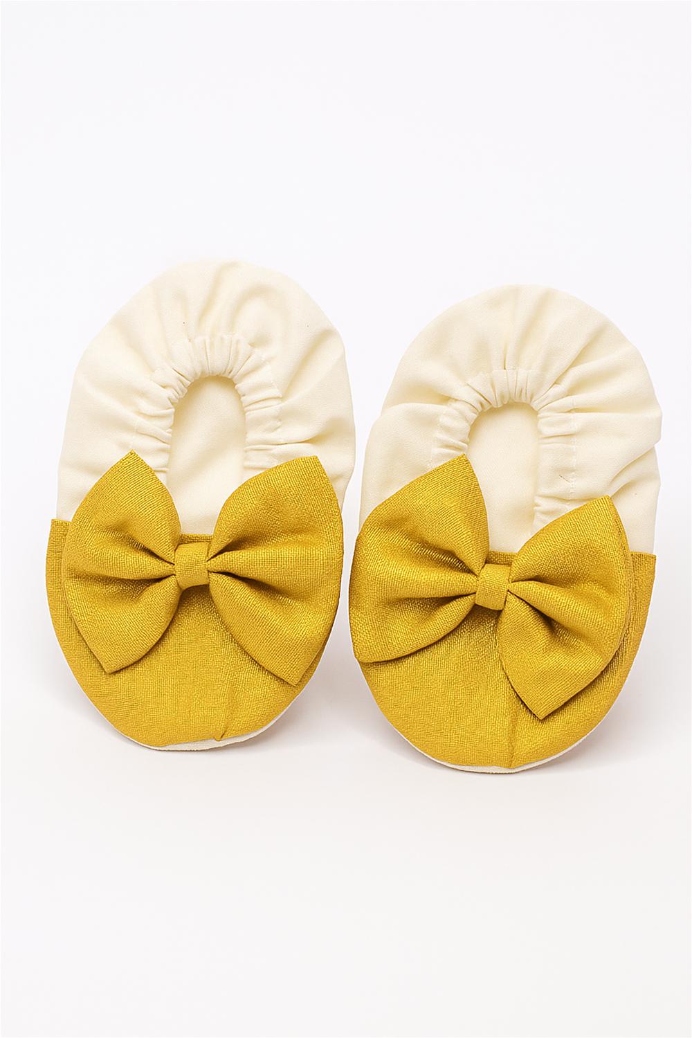A pair of off-white and golden baby booties with elasticated openings and large golden bows, displayed against a plain white background.