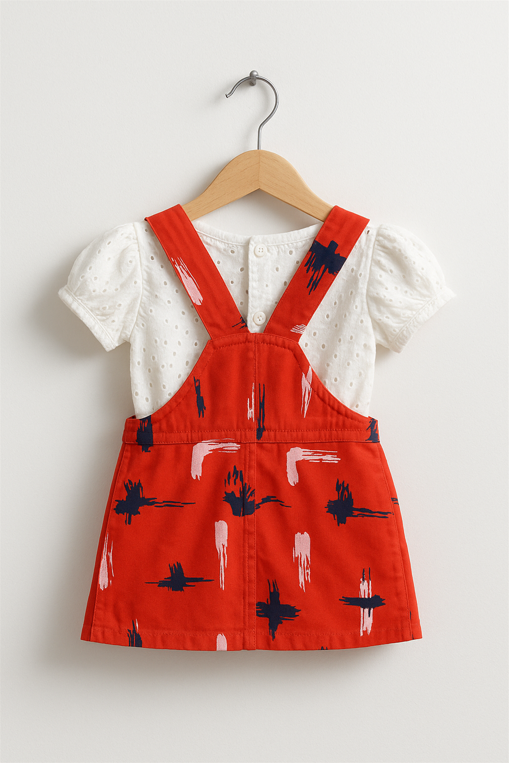 A back view of a baby girl's red pinafore dress with abstract brushstroke prints, displayed on a wooden hanger. The dress is paired with a white eyelet puff-sleeve inner top, showcasing crisscrossed straps and back button detailing