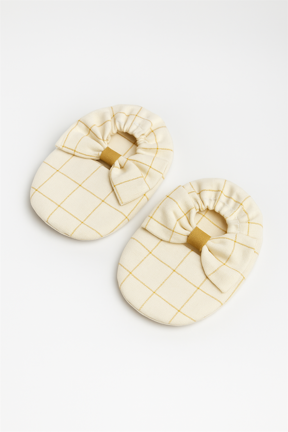 A pair of off-white baby booties with a mustard yellow grid pattern is neatly displayed on a white background. Each bootie is decorated with a soft bow at the front, adding a touch of charm.