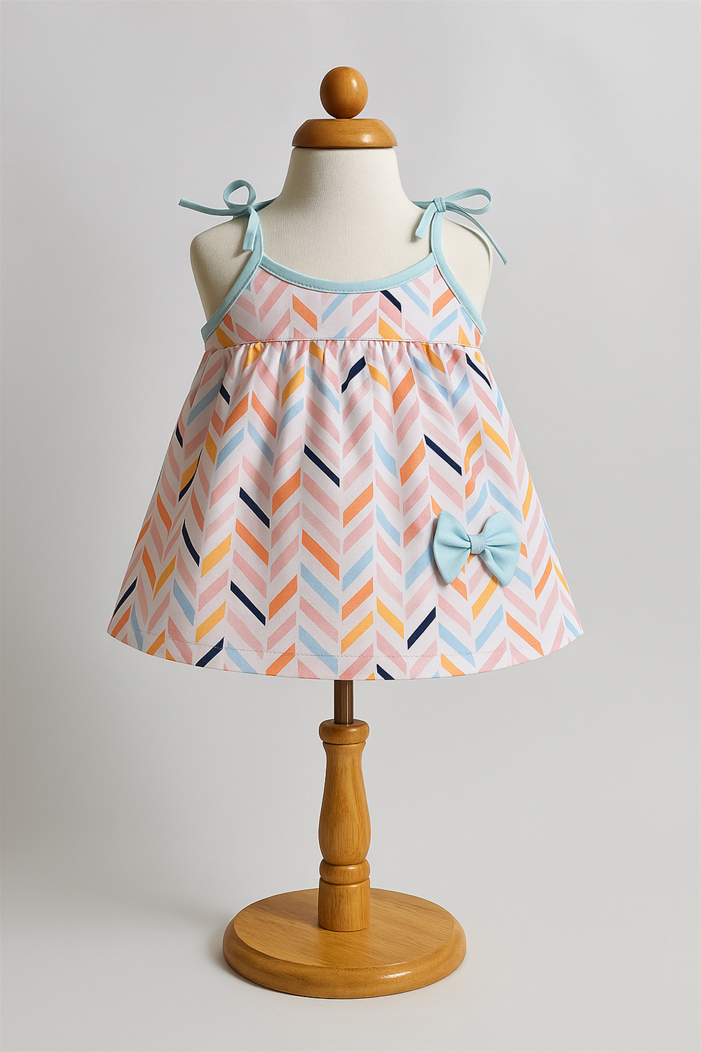 A baby dress with a vibrant pastel chevron pattern is displayed on a wooden stand mannequin. The dress features sky-blue shoulder ties, neckline piping, and a matching bow embellishment near the hem.