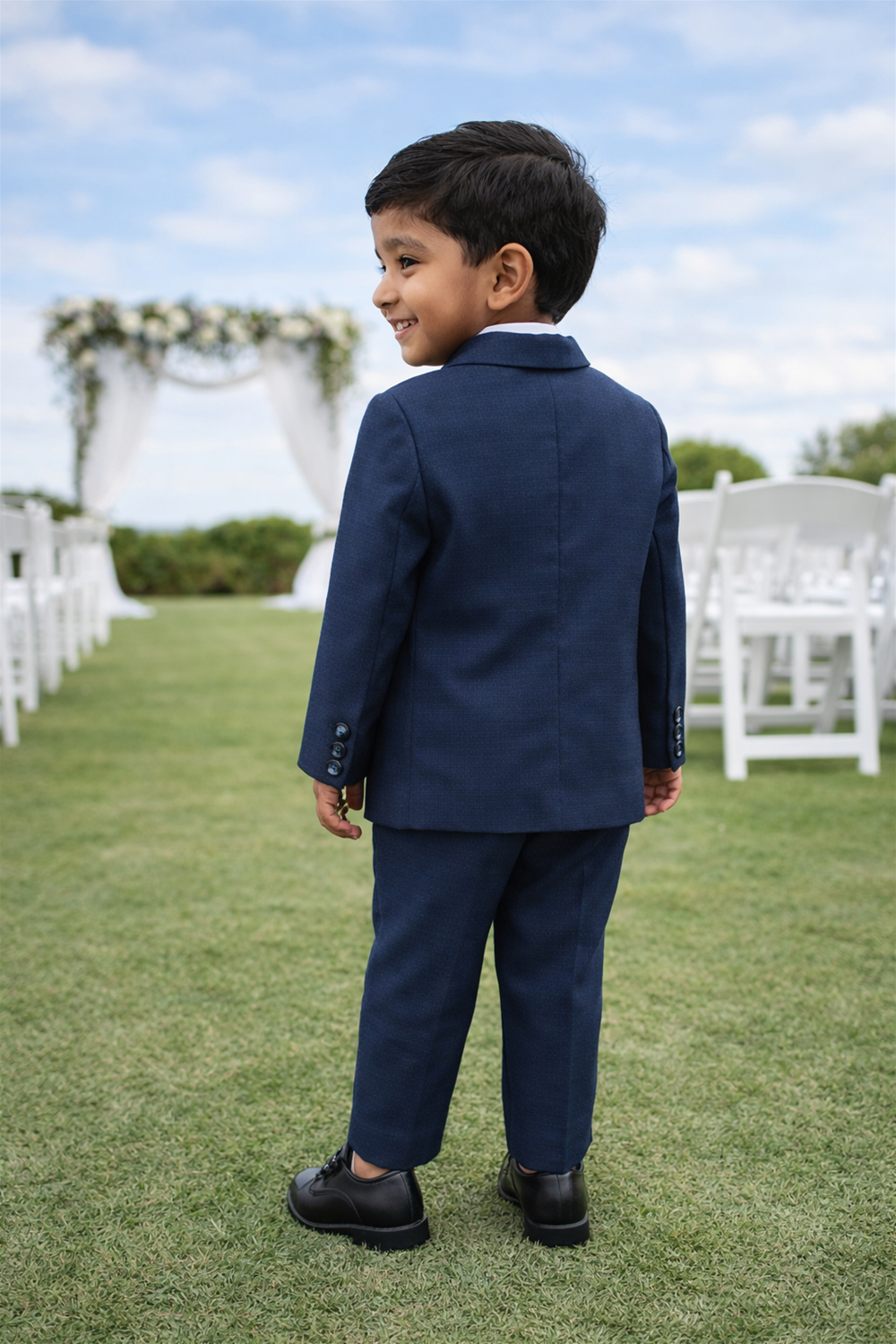 Toddler boy wearing formal suit walking outdoors on grass, seen from behind at a garden wedding ceremony setup.