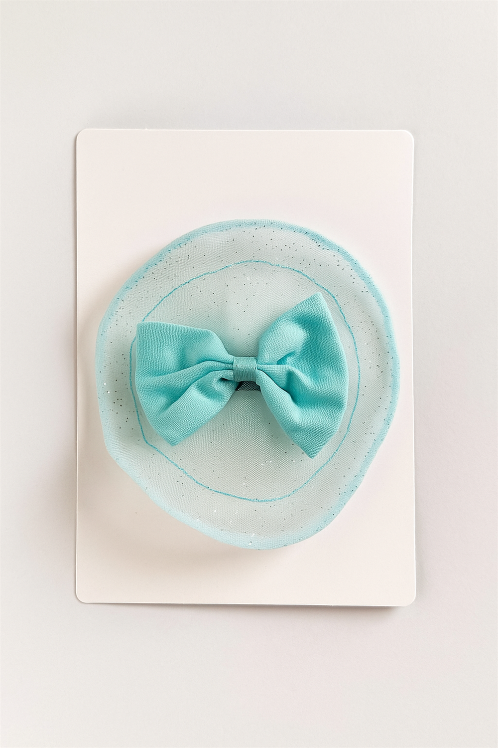 Aqua blue bow hair clip with sheer glittered circular base, displayed on a white card for elegant presentation.
