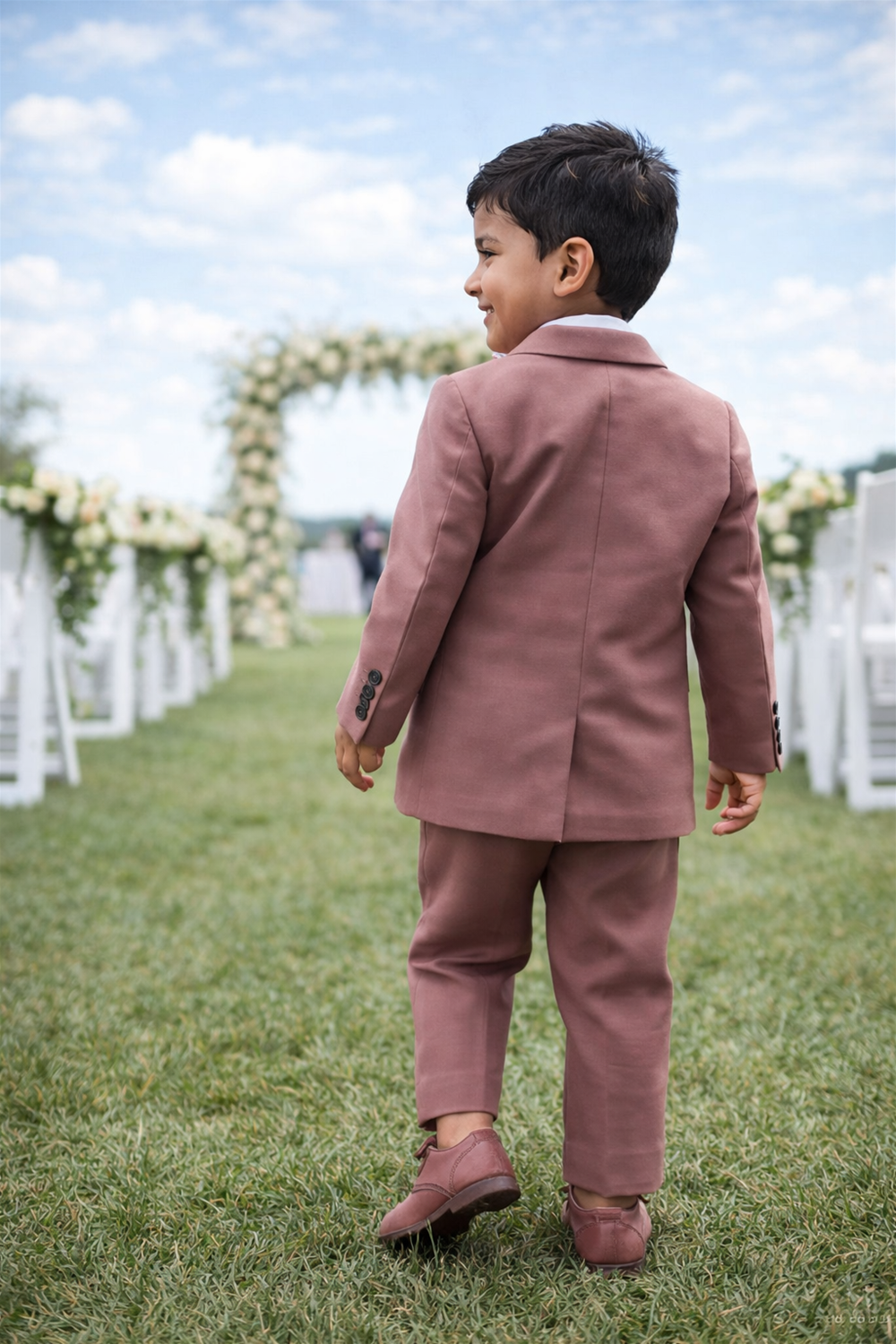 Toddler boy wearing formal suit walking outdoors on grass, seen from behind at a garden wedding ceremony setup.