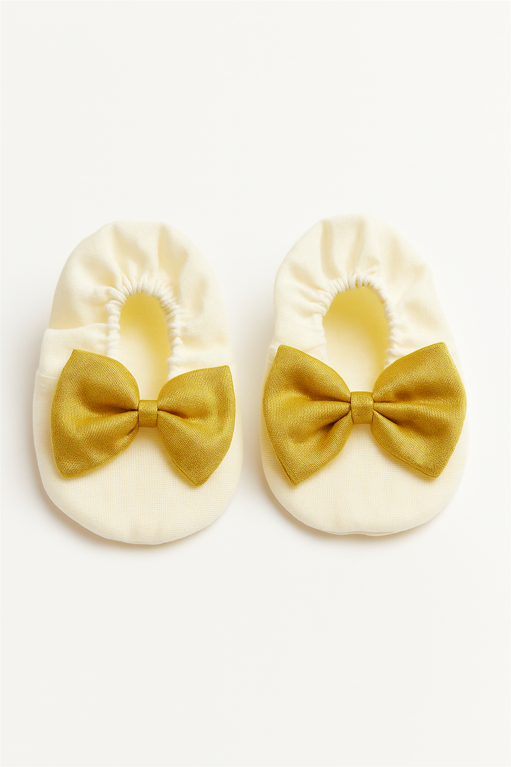 A pair of cream-colored baby booties adorned with golden bows is displayed against a white background. The soft fabric and elastic design highlight comfort and cuteness for infants.
