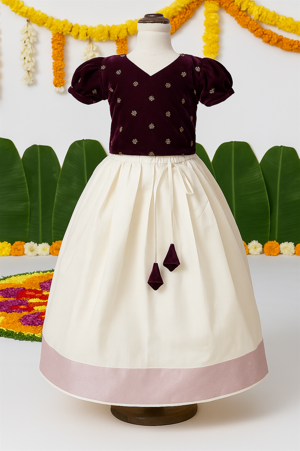 A traditional kids’ festive outfit is displayed on a mannequin, featuring a maroon embroidered blouse with puff sleeves and a cream long skirt with a pink border. The backdrop includes banana leaves and floral decorations, enhancing the Onam theme.