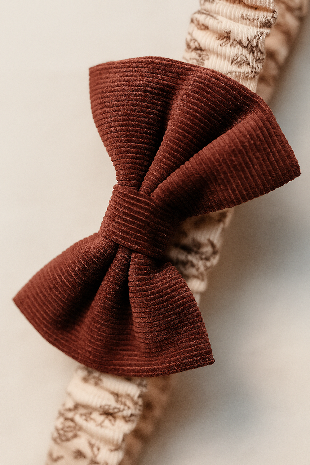 Close-up detail of textured burgundy corduroy bow attached to beige floral elastic baby headband, highlighting fabric texture and stitching.