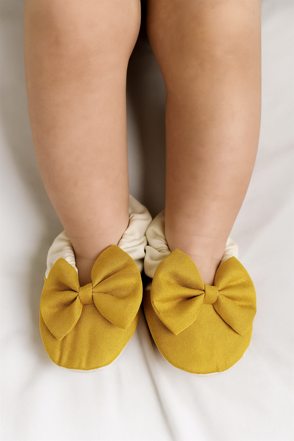 A baby wearing the same off-white and golden bow booties, with the shoes snugly fitted on the feet, photographed from above on a soft white surface.