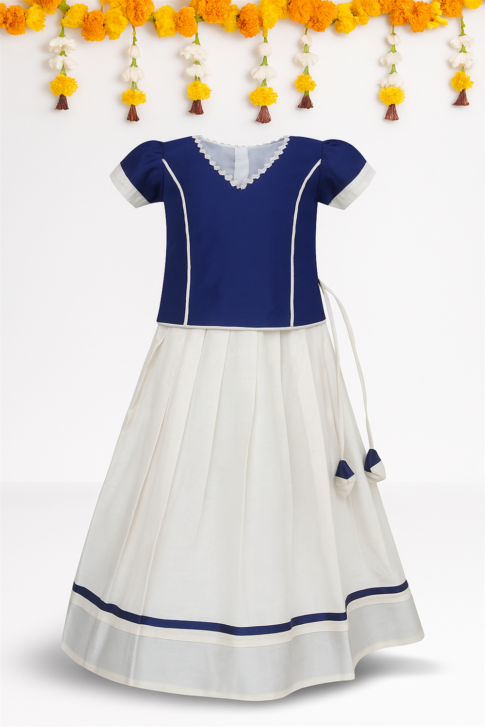 A traditional Kerala kidswear set displayed on a mannequin, featuring a navy blouse with white piping and a cream skirt with navy border. The outfit is adorned with tassels and showcased against a festive floral backdrop.