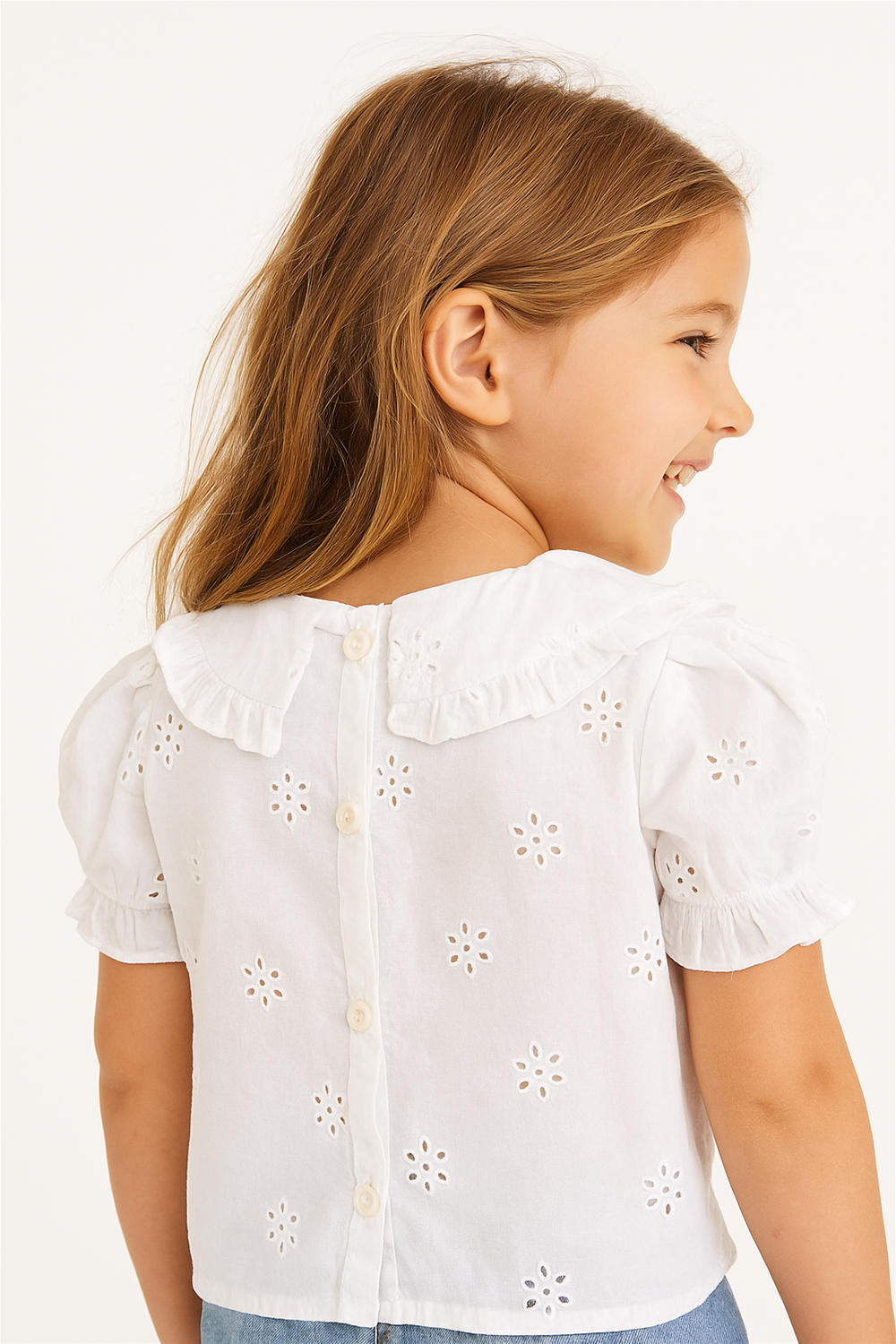 A young girl is shown from the back, smiling and looking to her right, wearing a white short-sleeve blouse with eyelet flower patterns and a ruffled collar. The blouse features a button-down back, and she pairs it with light blue denim jeans.