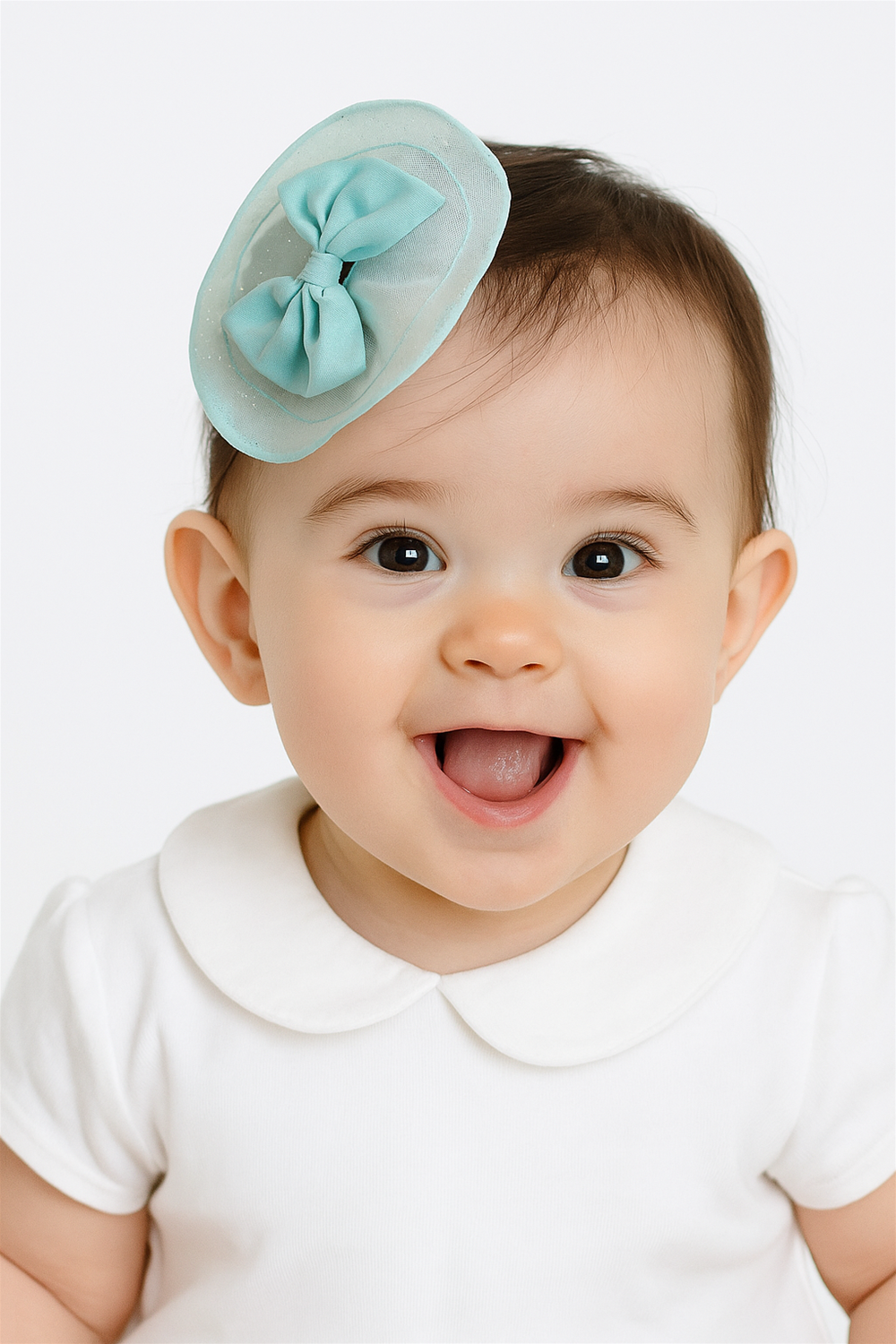 Happy baby girl wearing aqua blue bow hair clip on head, smiling brightly in a white dress with Peter Pan collar.