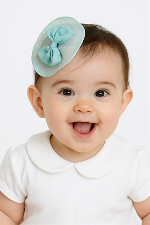 Happy baby girl wearing aqua blue bow hair clip on head, smiling brightly in a white dress with Peter Pan collar.