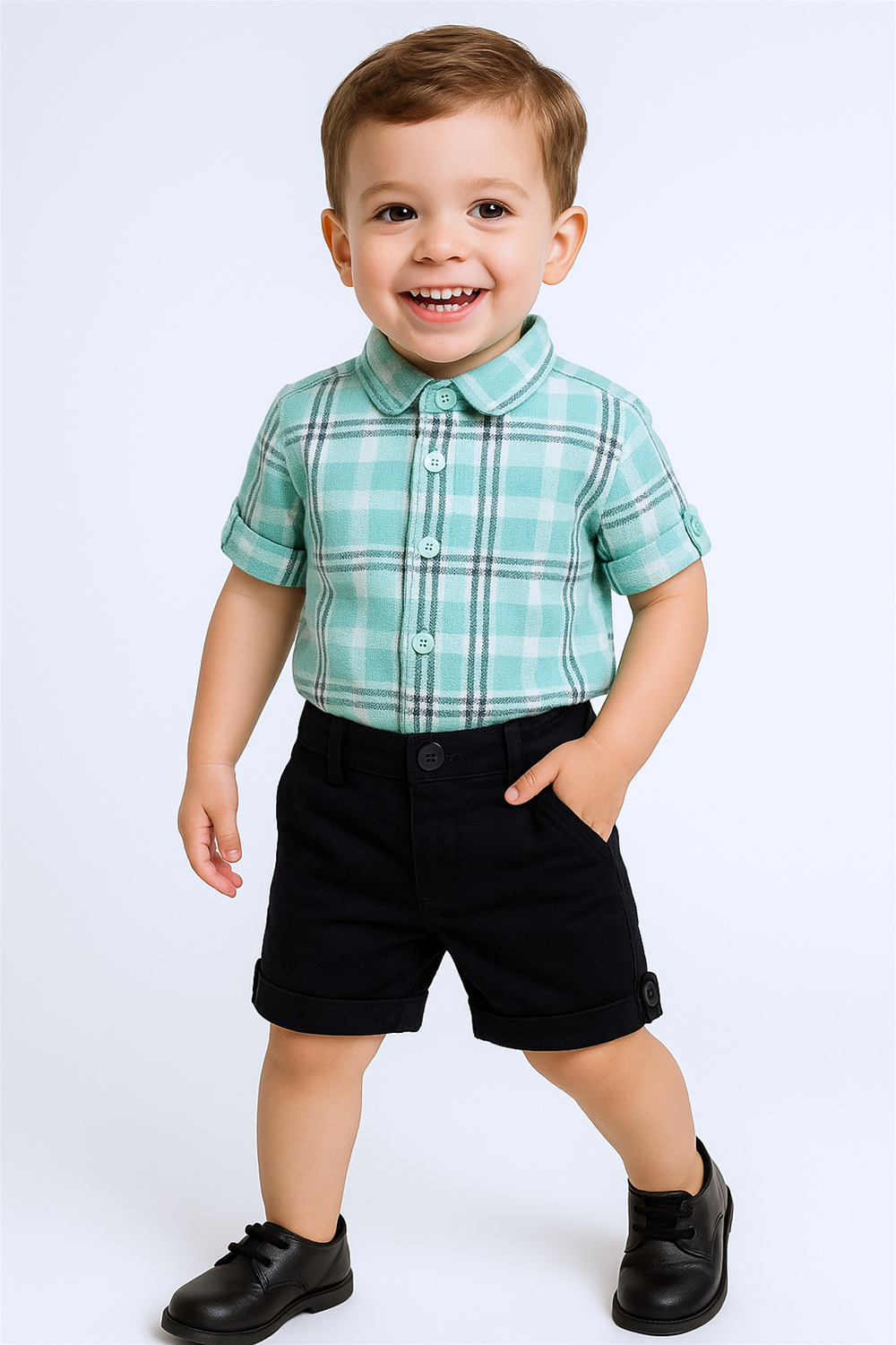 Smiling toddler boy wearing teal plaid shirt and black shorts, posing with hands in pockets against plain studio background.