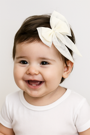 Smiling baby girl wearing cream bow hairclip with pearl accents on head, styled with white outfit, captured in natural lighting.