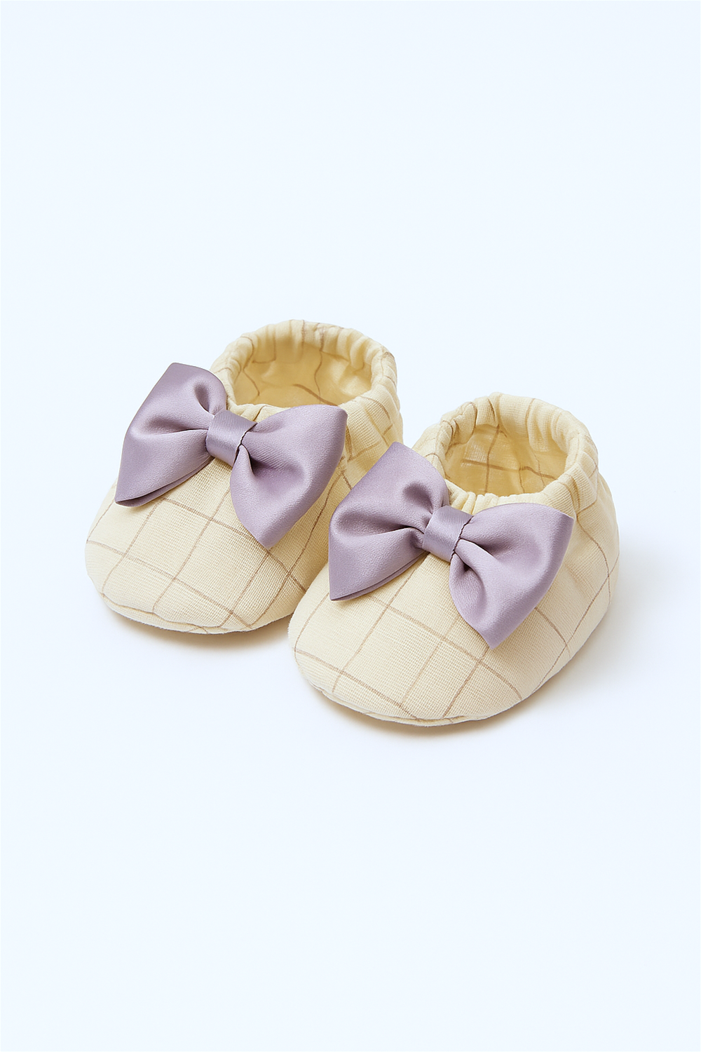 A pair of cream baby booties with a subtle checkered pattern, each adorned with a shiny lavender satin bow. The shoes are neatly displayed against a clean white background.