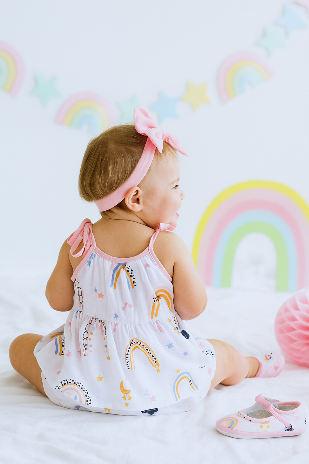 A baby girl is sitting on a white blanket, wearing a pastel rainbow-themed dress with tie straps and a matching pink bow headband. The scene is decorated with soft rainbow and star motifs, creating a dreamy and playful background.