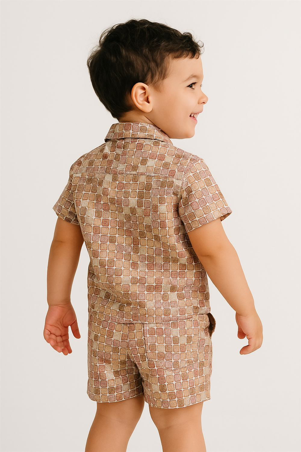 Cheerful toddler wearing beige patterned co-ord set, side view with hands relaxed, smiling naturally in bright studio lighting.