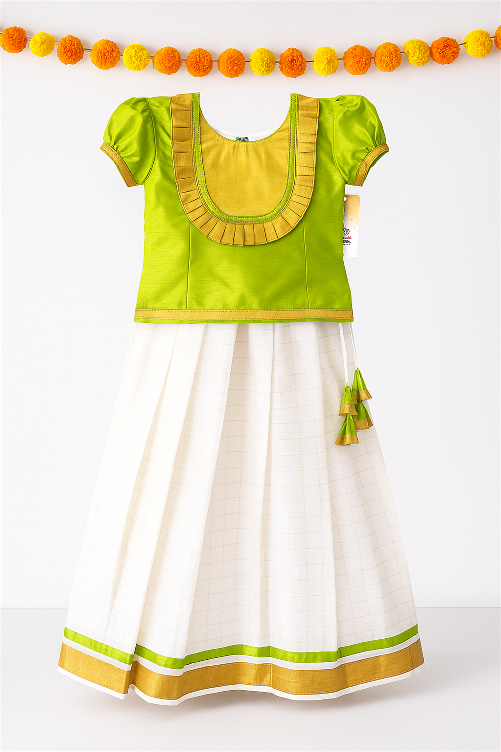 A beautifully displayed girls’ traditional Kerala skirt and top set with a green blouse featuring mustard pleated detailing and puff sleeves, paired with a cream skirt accented by golden and green borders. The outfit is photographed against a plain background with hanging orange and yellow marigold garlands for a festive touch.