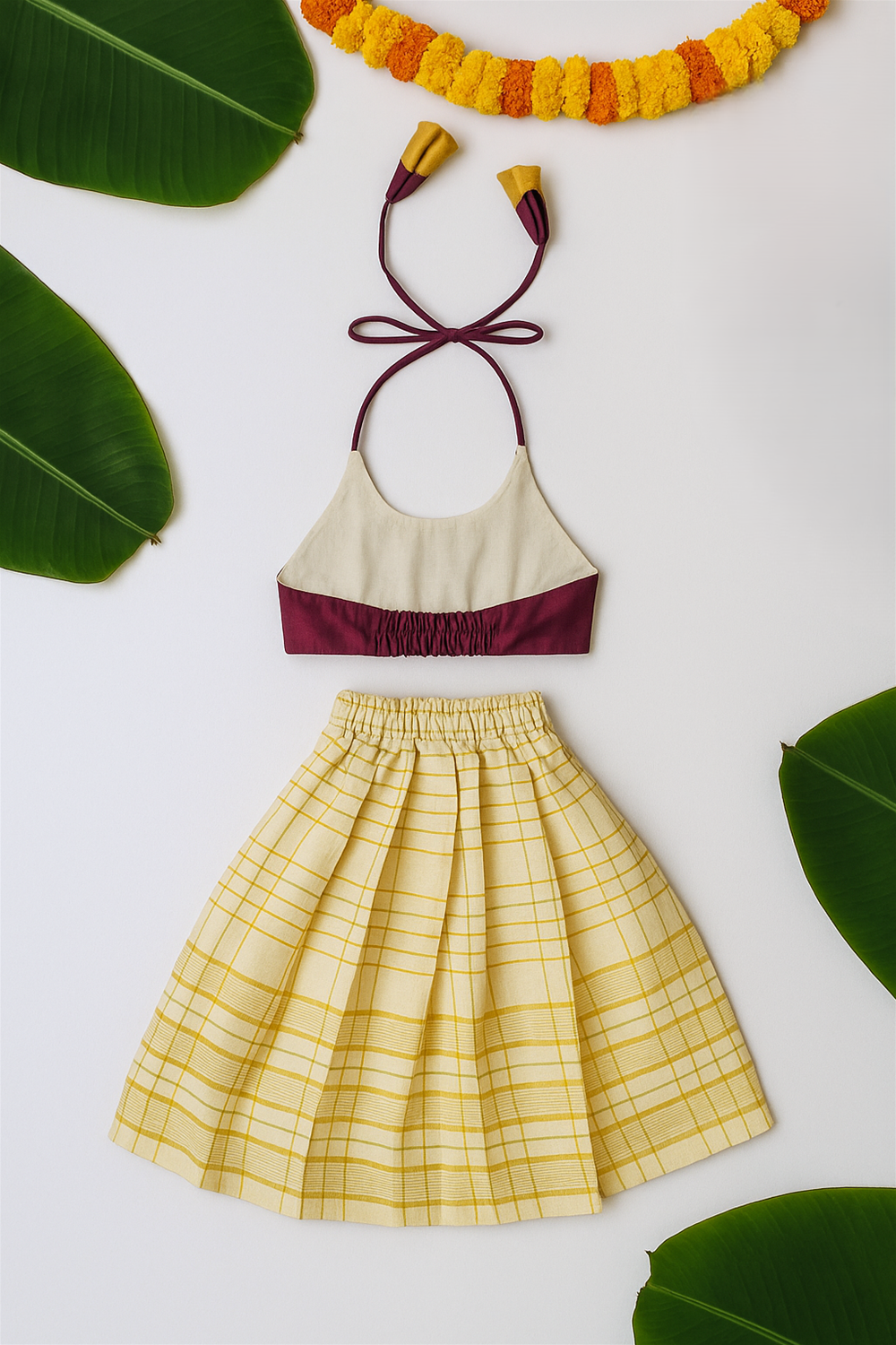 A flat lay of a festive kidswear set with a cream and maroon halter top and yellow checked skirt. Green banana leaves and orange garland decorate the white backdrop.