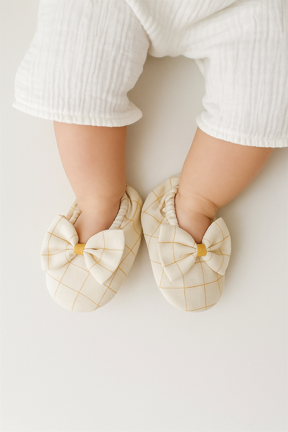 A baby is wearing the same off-white booties with a mustard yellow checkered design and a bow on top. The shoes fit snugly with elastic around the ankles, ensuring comfort and security.