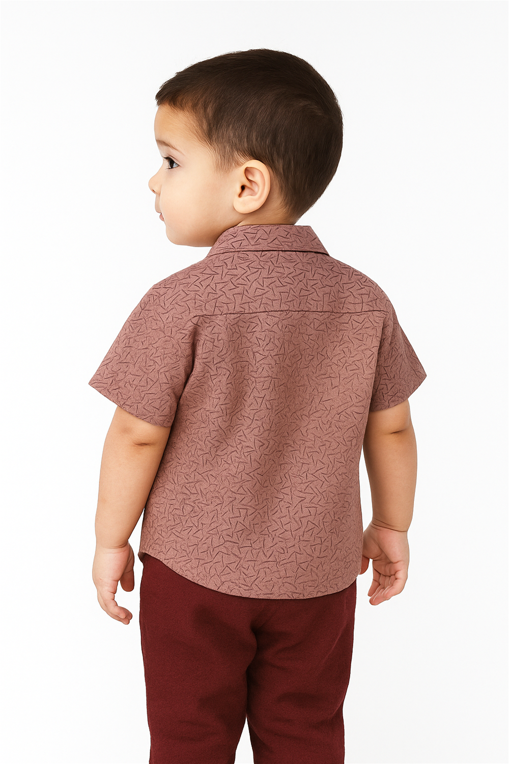 A young boy in a dull pink shirt stands against a wall, looking directly at the camera with a neutral expression.