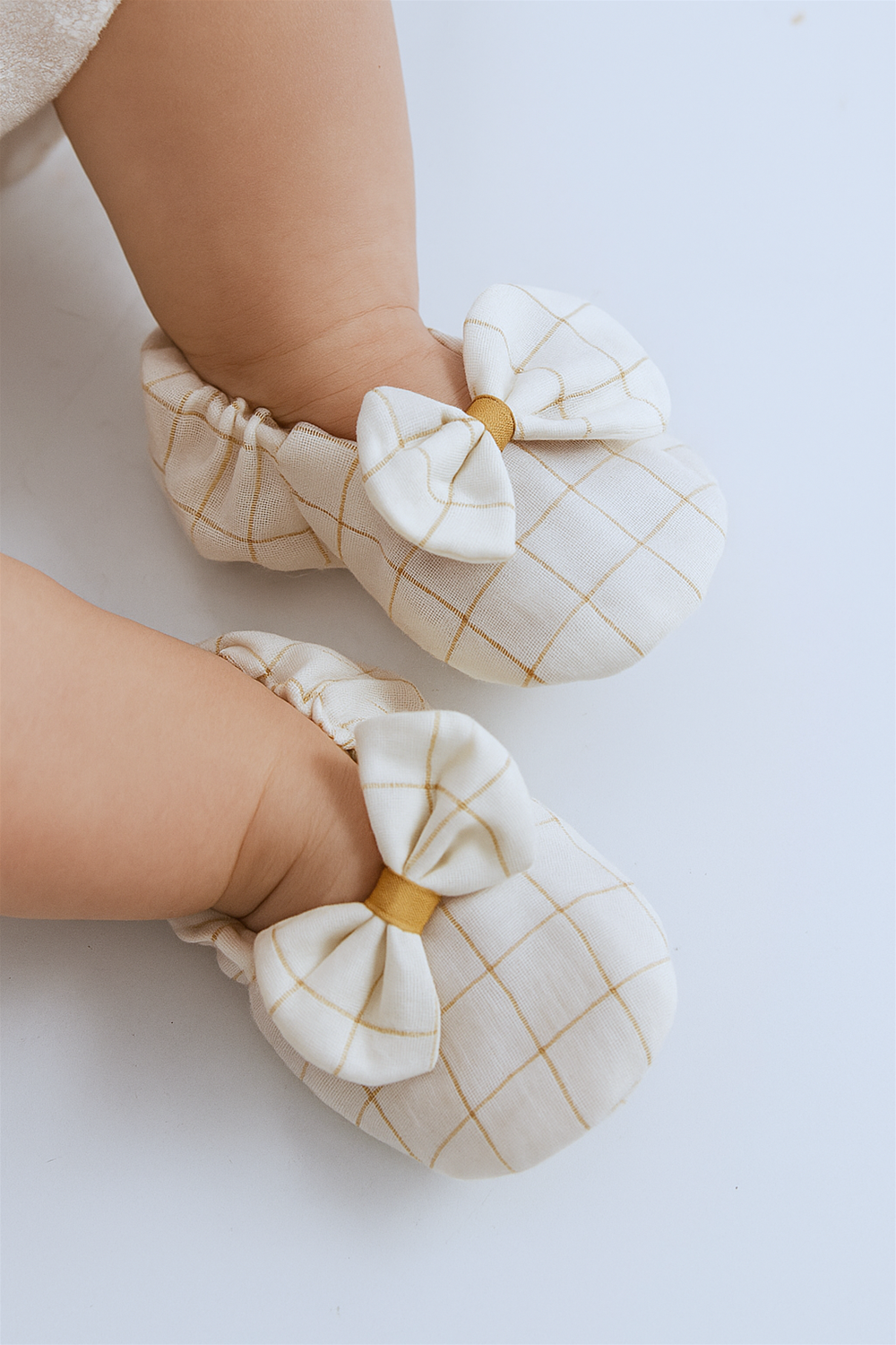 A baby is wearing the same off-white booties with a mustard yellow checkered design and a bow on top. The shoes fit snugly with elastic around the ankles, ensuring comfort and security.