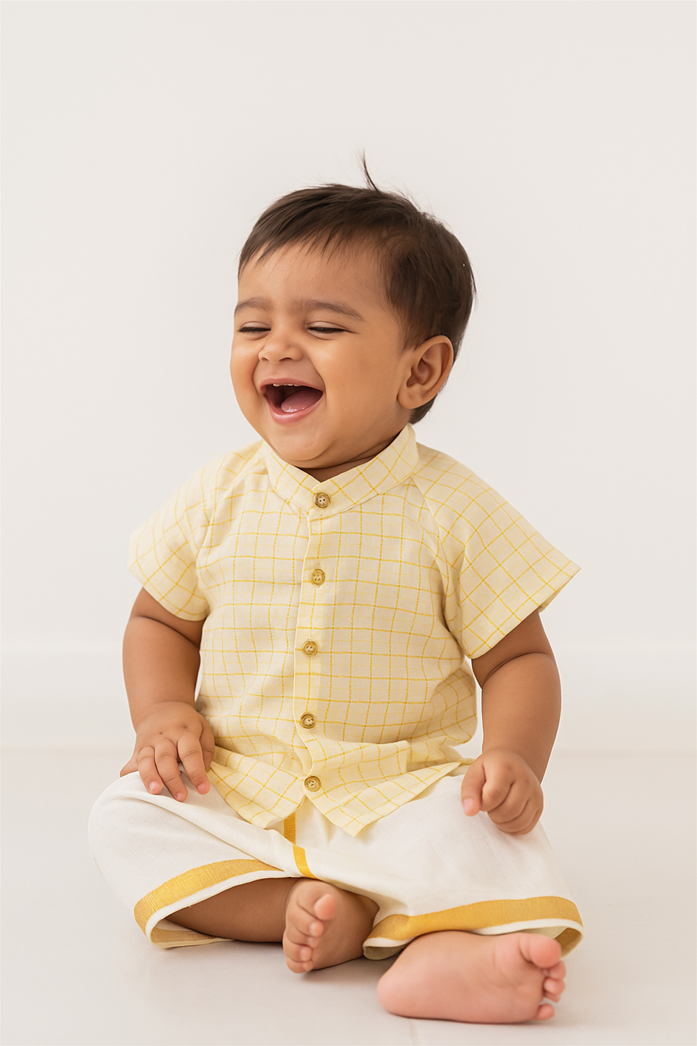 Smiling baby boy in yellow checked shirt and cream dhoti, sitting happily with bright expression, simple white background.
