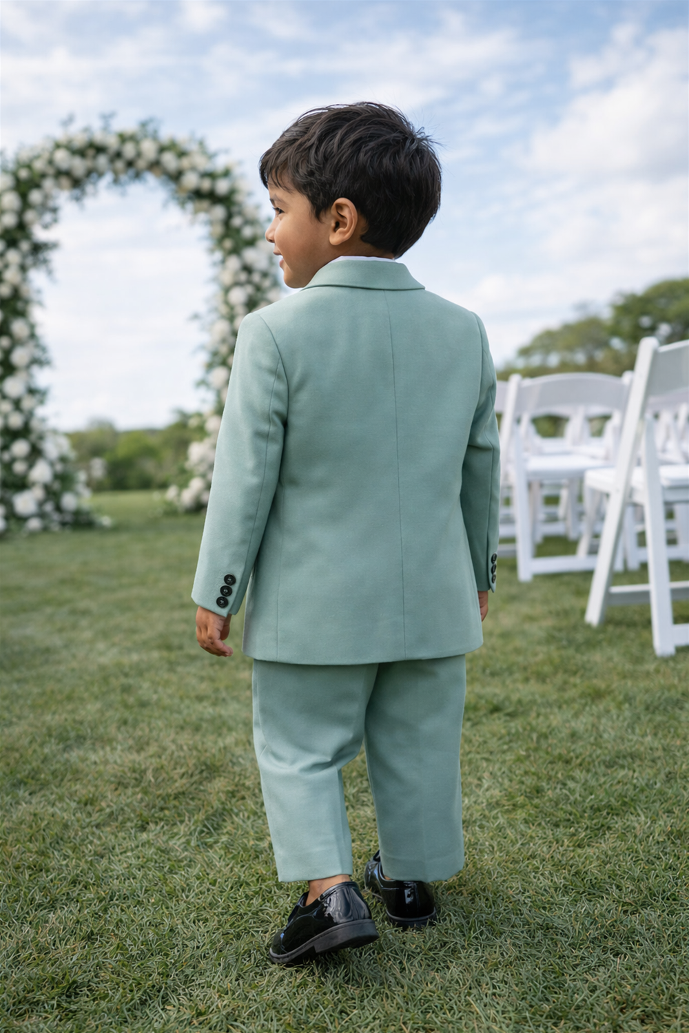 Toddler boy wearing mint green formal suit walking outdoors on grass, seen from behind at a garden wedding ceremony setup.