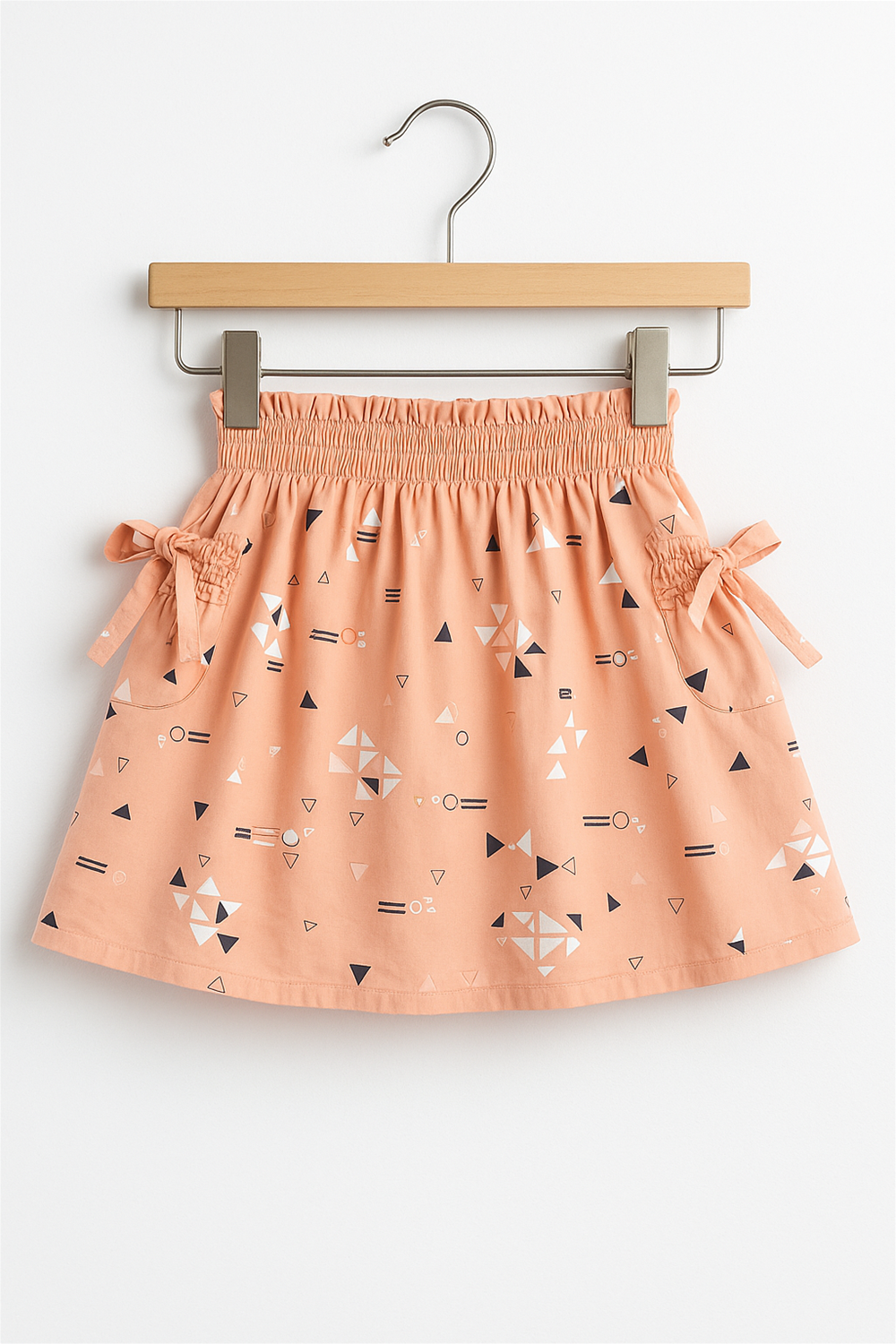 Peach-colored toddler skirt with elastic waist, side bows, and geometric print in white, black, and navy, displayed on hanger.