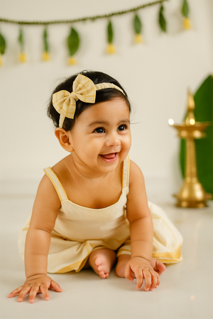 A smiling baby girl dressed in a cream and yellow frock with thin straps, wearing a matching yellow checkered bow hairband. She is sitting on the floor with a festive background featuring a brass lamp and green leaf decorations.