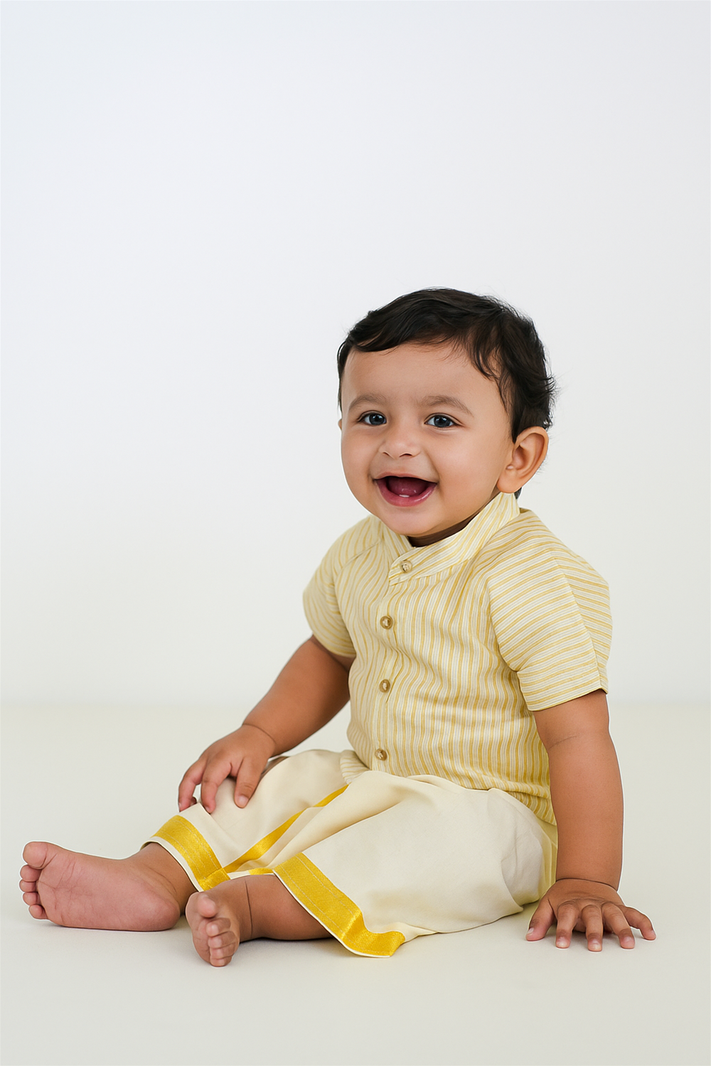 Cheerful baby boy in yellow striped kurta and traditional white dhoti, sitting indoors on plain background, looking happy and joyful.