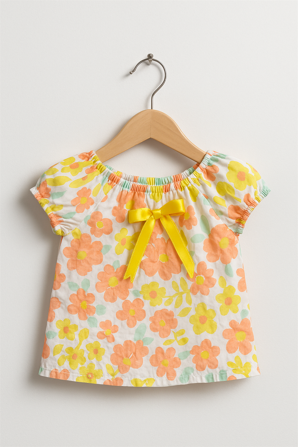 Floral baby girl’s top with bright yellow bow at neckline, short puff sleeves, displayed on wooden hanger, cheerful vibrant design.